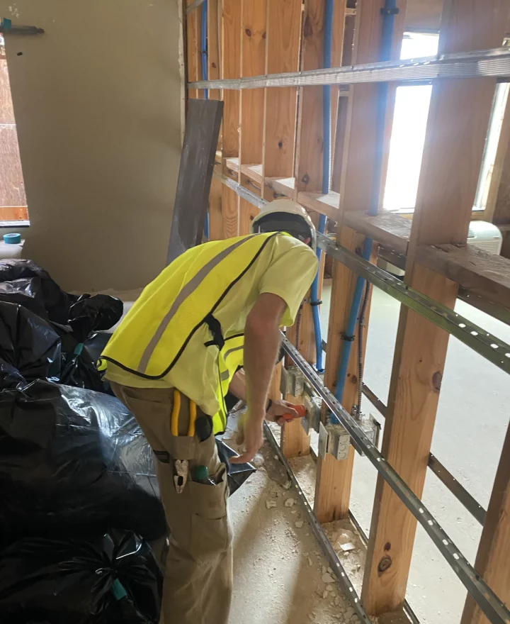 An electrician in safety gear installs electrical components within an open wall framework.