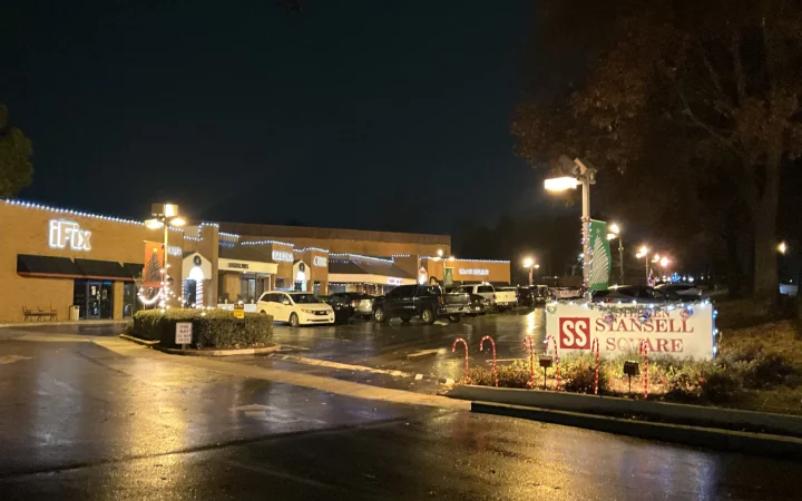 A nighttime view of a commercial plaza with festive lighting and decorations, showcasing businesses like 'eFix' and a holiday sign for 'Stansell Square.'