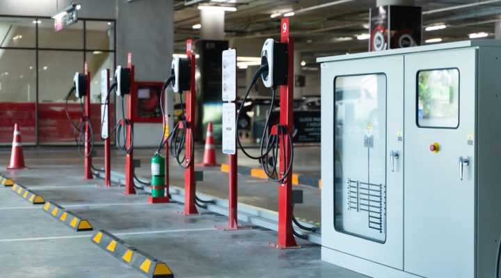Multiple red EV charging stations lined up in an indoor parking garage, with safety cones and a large power distribution cabinet nearby.