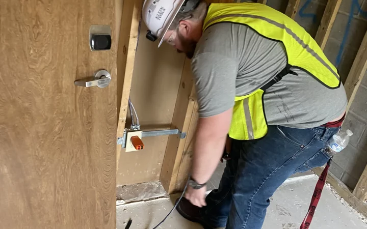 An electrician in a hard hat and reflective vest works on electrical wiring in a construction area.