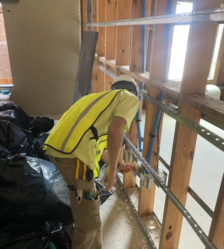 An electrician in safety gear installs electrical components within an open wall framework.