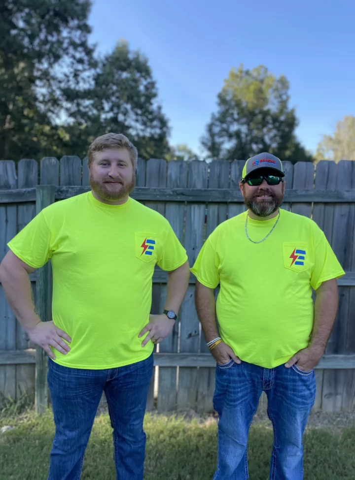 Two electrical service technicians in bright yellow shirts and jeans standing in front of a wooden fence.